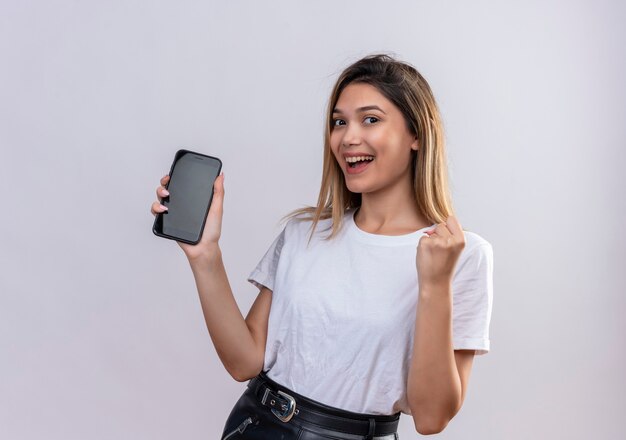 Une belle jeune femme en t-shirt blanc souriant tout en montrant l'espace vide du téléphone mobile avec le poing fermé sur un mur blanc