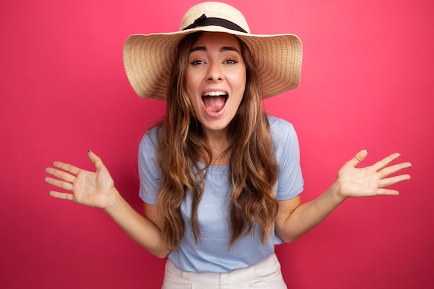 Belle jeune femme en t-shirt bleu et chapeau d'été regardant la caméra follement heureuse et excitée en levant les bras debout sur fond rose