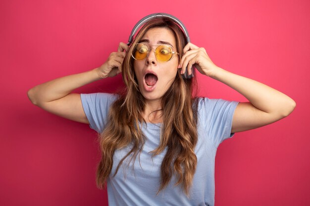 Belle jeune femme en t-shirt bleu portant des lunettes jaunes avec des écouteurs à côté étonné et surpris