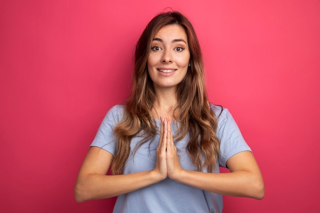 Belle Jeune Femme En T-shirt Bleu Regardant La Caméra Avec Le Sourire Sur Le Visage, Main Dans La Main Comme Un Geste De Namaste Debout Sur Fond Rose