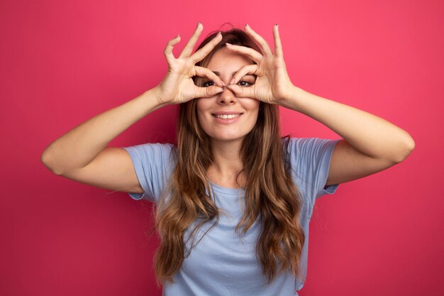 Belle jeune femme en t-shirt bleu regardant à travers les doigts faisant un geste binoculaire souriant joyeusement