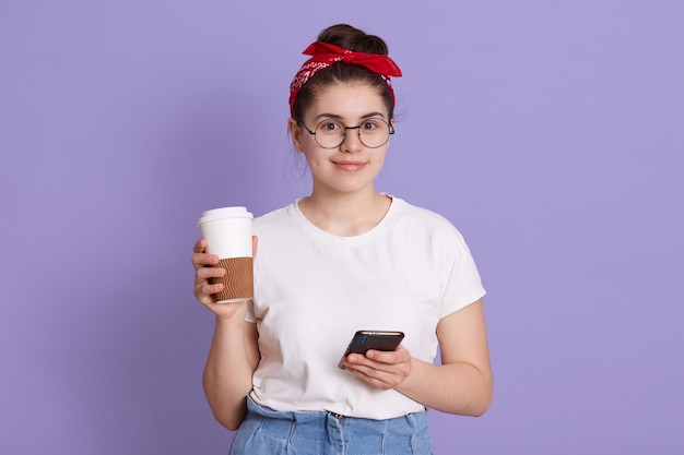 Belle jeune femme avec téléphone intelligent et café à emporter, fille étudiante souriante posant isolé sur espace lilas