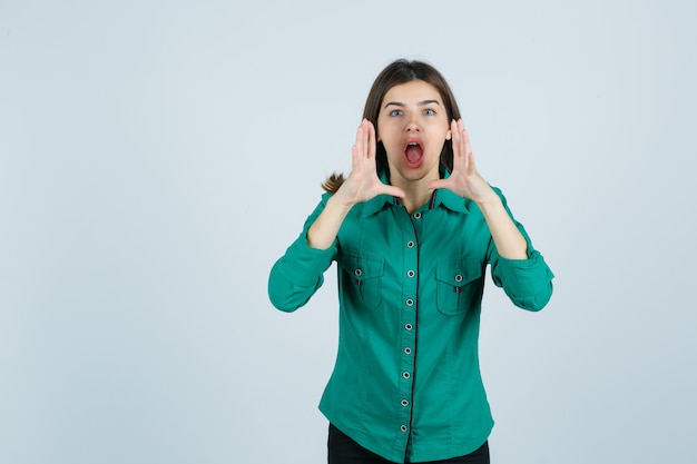 Belle jeune femme tenant la main près de la bouche tout en criant en chemise verte et à la vue excitée, de face.