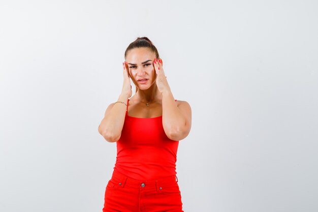 Belle jeune femme tenant les mains sur les tempes en débardeur rouge, pantalon et à la fatigue. vue de face.