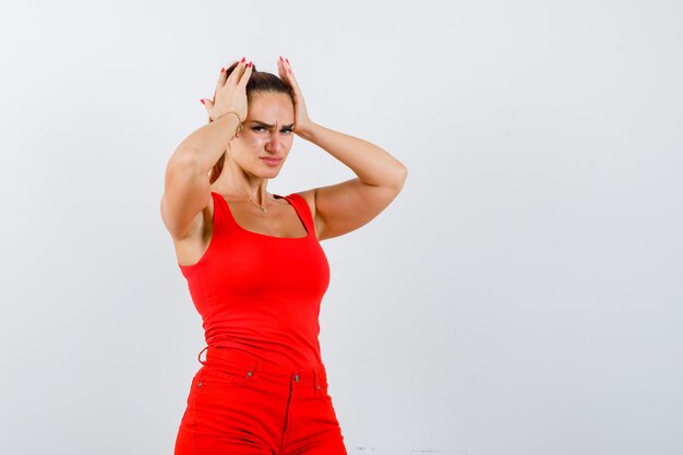 Belle jeune femme tenant les mains sur la tête en débardeur rouge, pantalon et regardant déprimé, vue de face.