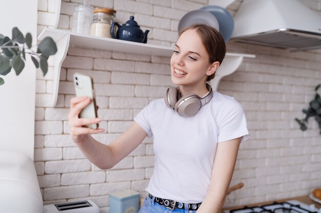 Belle jeune femme en tenue décontractée est assise sur la table de la cuisine avec des écouteurs, faisant un appel vidéo