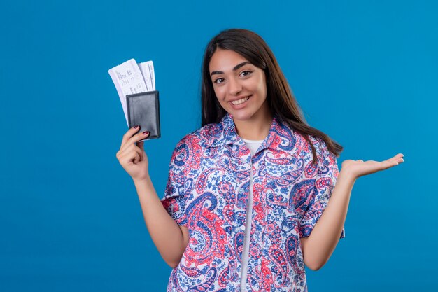 Belle jeune femme touriste tenant un passeport avec des billets souriant joyeusement, présentant le bras de la main, prêt à partir en vacances sur mur bleu isolé