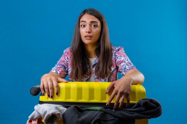 Belle jeune femme touriste avec une valise pleine de vêtements ayant des doutes et avec l'expression du visage confus sur mur bleu isolé