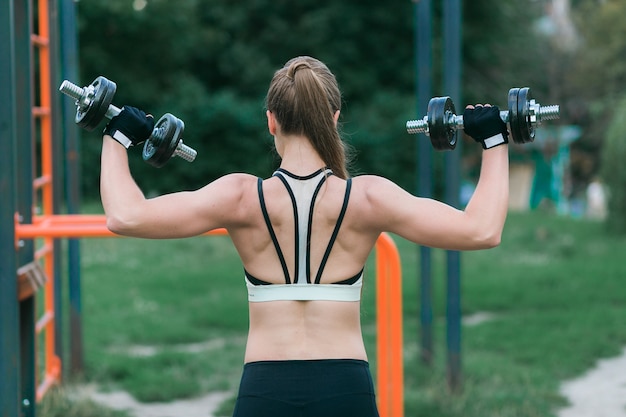 Belle jeune femme travaille sur ses bras, son dos et ses épaules avec des haltères à l&#39;extérieur