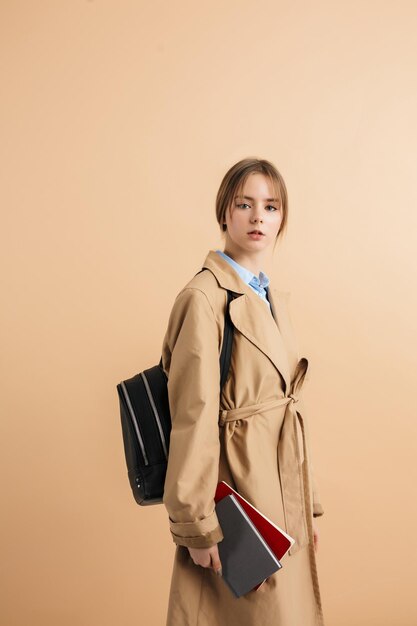 Belle jeune femme en trench-coat avec sac à dos sur l'épaule tenant des blocs-notes à la main regardant pensivement à huis clos sur fond beige isolé