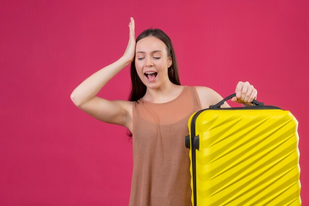 Belle jeune femme avec valise de voyage debout avec la main sur la tête pour erreur se souvenir de l'erreur sur fond rose