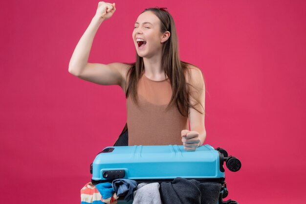 Belle jeune femme avec valise de voyage levant le poing après une victoire sortie et heureux sur mur rose