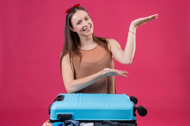 Belle jeune femme avec valise de voyage présentant avec bras de mains souriant joyeusement sur mur rose
