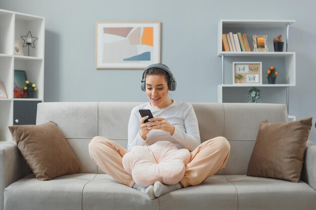 Belle jeune femme en vêtements décontractés avec un casque assis sur un canapé à l'intérieur de la maison tenant un smartphone heureux et surpris en regardant l'écran de son mobile souriant joyeusement relaxant