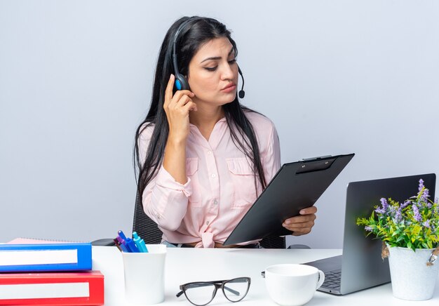 Belle jeune femme en vêtements décontractés avec un casque et un microphone tenant un presse-papiers le regardant avec un visage sérieux assis à la table avec un ordinateur portable sur blanc