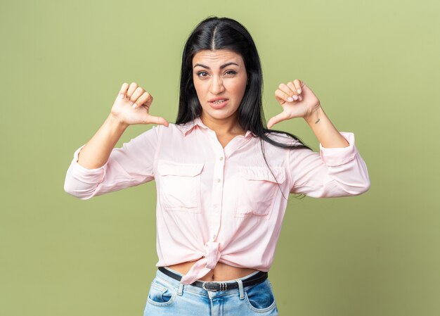 Belle jeune femme en vêtements décontractés confus se pointant sur elle-même debout sur un mur vert