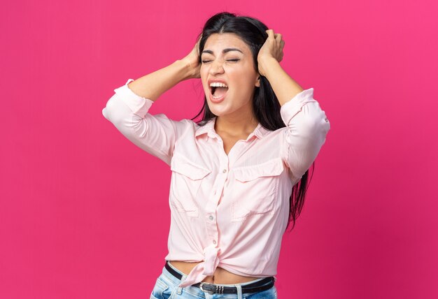 Belle jeune femme en vêtements décontractés criant et hurlant d'être frustré d'être frustré en tirant ses cheveux debout sur le rose