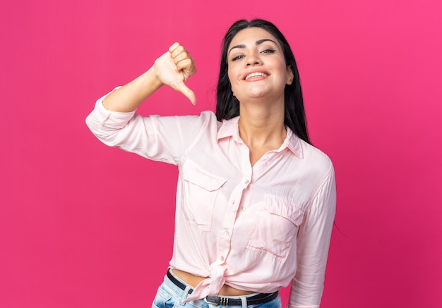 Belle jeune femme en vêtements décontractés, heureuse et positive souriante montrant joyeusement les pouces vers le bas, debout sur le mur rose