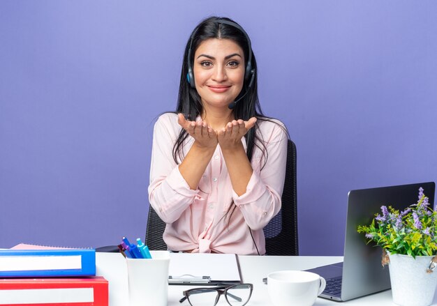 Belle jeune femme en vêtements décontractés portant un casque avec microphone heureux et positif se tenant la main devant elle assise à la table avec un ordinateur portable sur un mur bleu travaillant au bureau