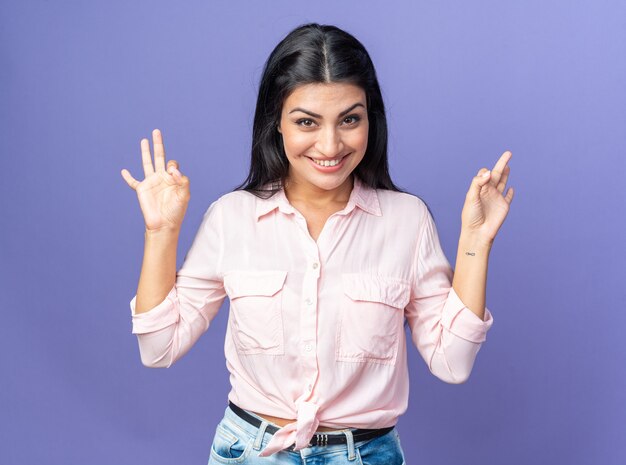 Belle jeune femme en vêtements décontractés regardant devant souriant joyeusement faisant signe ok debout sur le mur bleu