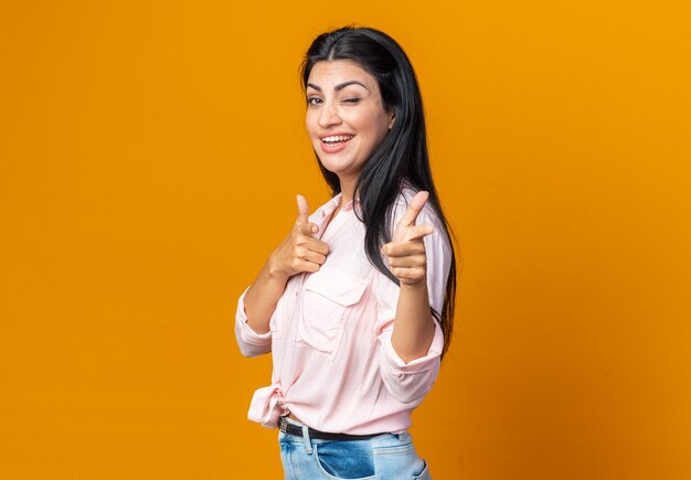 Belle jeune femme en vêtements décontractés souriante confiante, heureuse et joyeuse, pointant avec l'index à l'avant, debout sur le mur orange