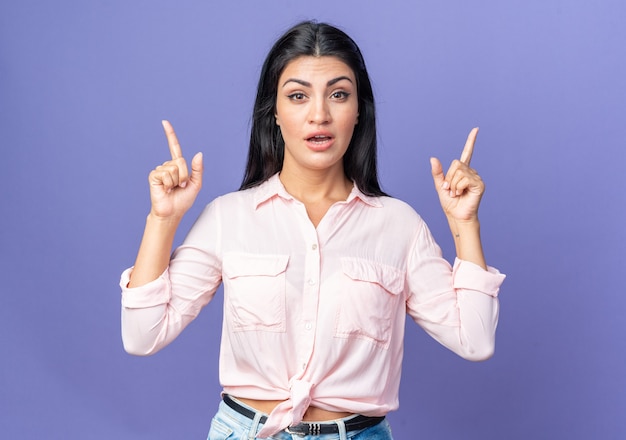 Belle jeune femme en vêtements décontractés surpris pointant avec l'index debout sur le mur bleu