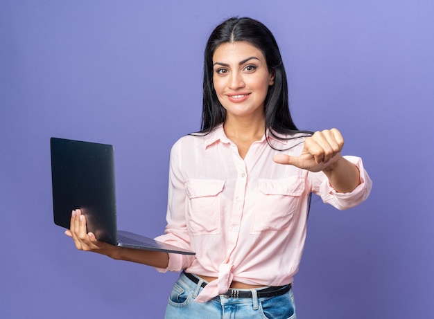 Belle jeune femme en vêtements décontractés tenant un ordinateur portable à la recherche de sourire pointant avec le pouce sur un ordinateur portable