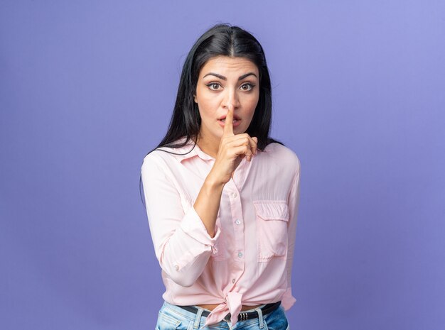 Belle jeune femme en vêtements décontractés avec un visage sérieux faisant un geste de silence avec le doigt sur les lèvres debout sur le mur bleu