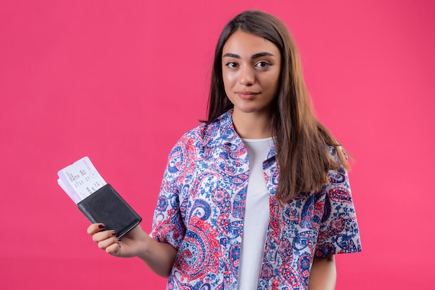Belle jeune femme de voyageur tenant un passeport et des billets regardant la caméra avec une expression confiante sérieuse debout sur fond rose isolé