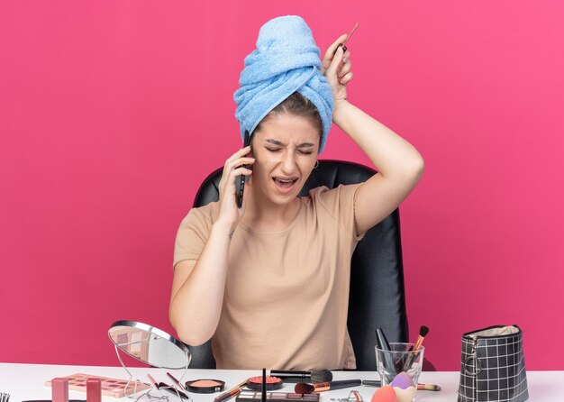 Une belle jeune fille agacée est assise à table avec des outils de maquillage enveloppés de cheveux dans une serviette tenant un brillant à lèvres parle au téléphone isolé sur un mur rose