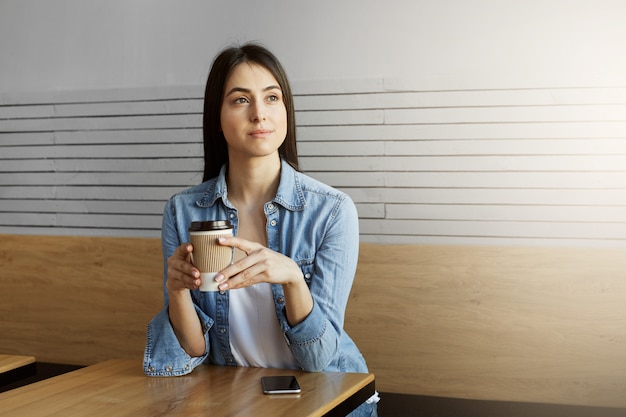 Belle jeune fille aux cheveux noirs en chemise en jean et t-shirt blanc boit du café, regardant de côté avec une expression détendue et attendant un ami en retard.