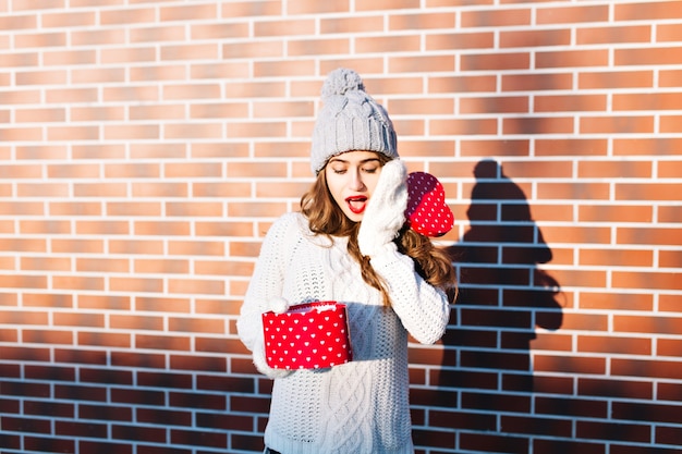 Belle jeune fille en bonnet tricoté et gants sur le mur extérieur. Elle tient un cadeau ouvert dans ses mains, a l'air surprise.