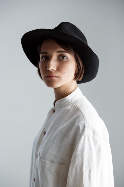 Belle jeune fille brune au chapeau noir sur mur blanc