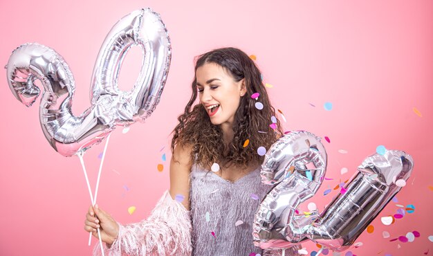 Belle jeune fille brune aux cheveux bouclés posant sur un fond de studio rose avec des confettis et tenant dans sa main des ballons d'argent pour le concept de nouvel an