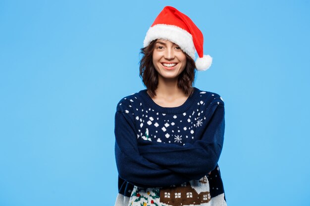 Belle jeune fille brune en pull en tricot et chapeau de Noël souriant sur mur bleu