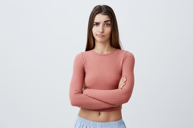 Belle jeune fille brune de race blanche malheureuse avec une longue coiffure en tenue à la mode, croisant les mains avec une expression sarcastique