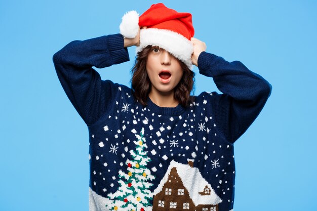 Belle jeune fille brune surprise en pull en tricot et chapeau de Noël sur mur bleu