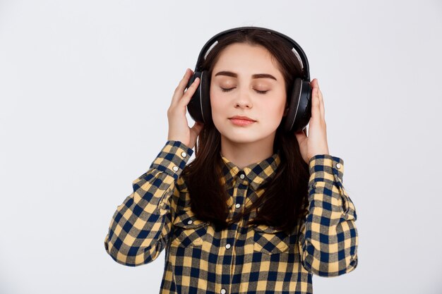 Belle jeune fille brune vêtue d'une chemise à carreaux enjoing musique dans les écouteurs avec les yeux fermés sur le mur blanc