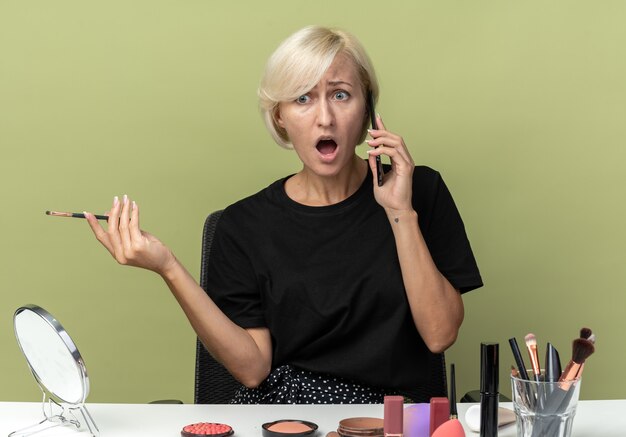 Une belle jeune fille en colère est assise à table avec des outils de maquillage parle au téléphone tenant un pinceau de maquillage isolé sur un mur vert olive