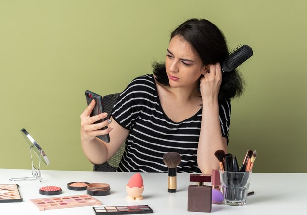 Une belle jeune fille confuse est assise à table avec des outils de maquillage tenant un peigne et regardant le téléphone dans sa main isolée sur un mur vert olive