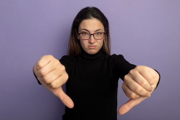 Photo gratuite belle jeune fille dans un col roulé noir et des lunettes d'être mécontent montrant les pouces vers le bas