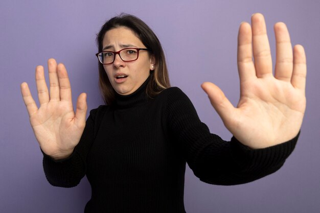 Belle jeune fille dans un col roulé noir et des lunettes faisant le geste d'arrêt tenant la main ouverte d'avoir peur