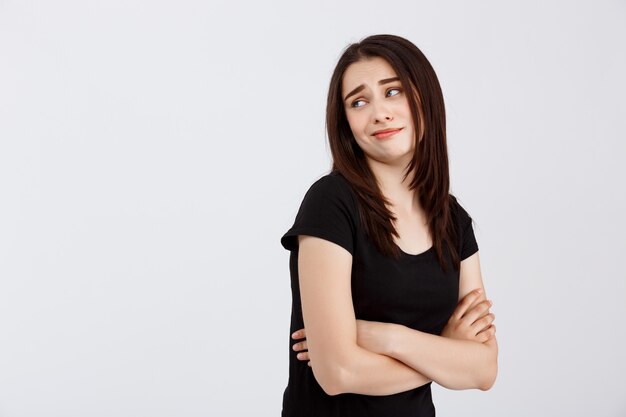 Belle jeune fille déçue en t-shirt noir posant sur un mur blanc