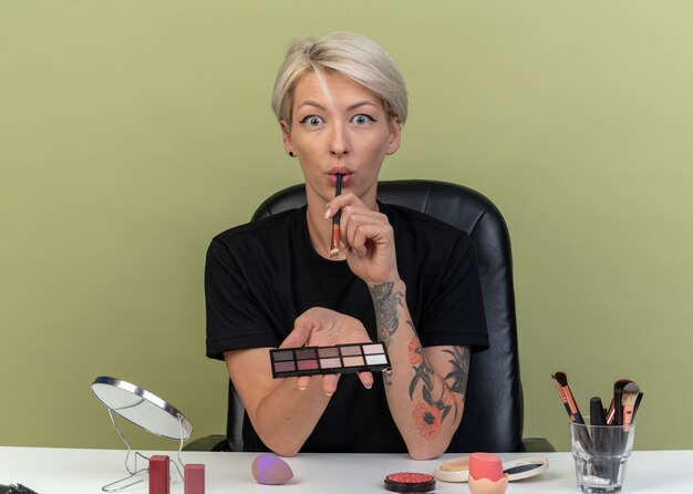 Photo gratuite une belle jeune fille effrayée est assise à table avec des outils de maquillage tenant une palette de fards à paupières avec un pinceau de maquillage isolé sur fond vert olive