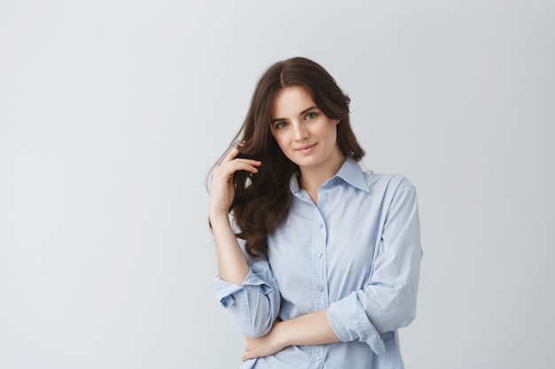 Belle jeune fille étudiante en chemise bleue tenant la main dans de longs cheveux ondulés posant pour un article de journal étudiant sur les meilleurs étudiants de l'année.