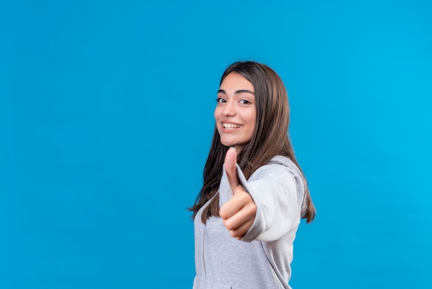 Belle jeune fille en gris à capuche regardant la caméra avec le sourire sur le visage faisant comme un geste à la caméra debout sur fond bleu