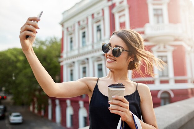 Belle jeune fille hispanique brune aux lunettes de soleil une robe noire souriante avec des dents, prenant selphie devant un beau bâtiment rouge, boire du café, passer du bon temps après la boutique