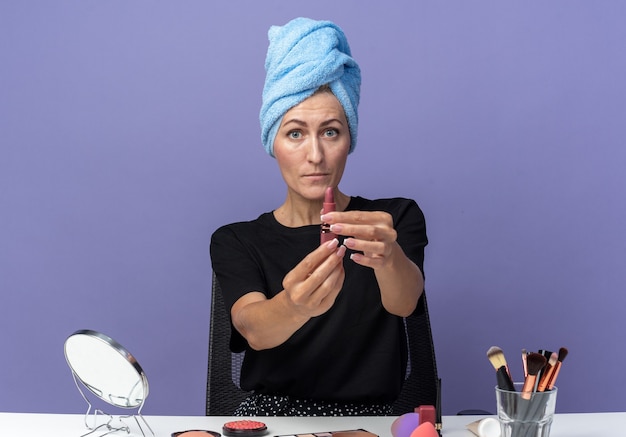 Photo gratuite une belle jeune fille inquiète est assise à table avec des outils de maquillage essuyant les cheveux dans une serviette tenant du rouge à lèvres à la caméra isolée sur un mur bleu