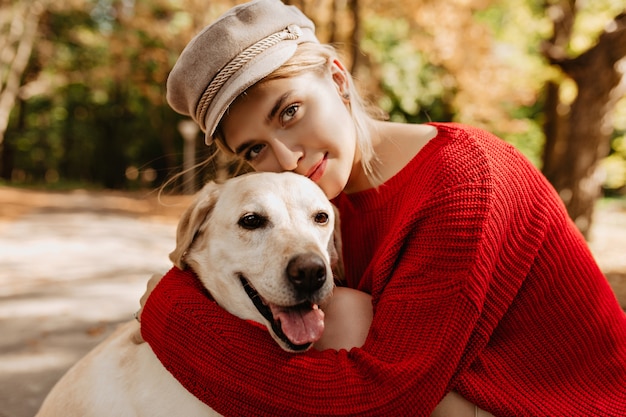 Belle jeune fille en joli pull rouge à la mode étreignant le labrador dans la forêt. Jolie blonde au chapeau léger avec son chien assis dans le parc.