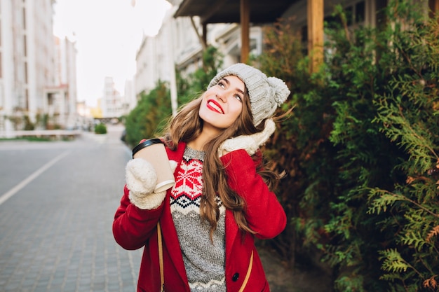 Belle jeune fille en manteau rouge et bonnet tricoté marchant dans la rue. Elle tient le café pour aller dans des gants blancs, rêvant au ciel.